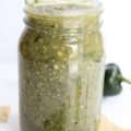 Clear jar holding green salsa. Pictures of jalapeños behind jar.