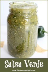 Jar with green salsa, peppers and chips behind it.